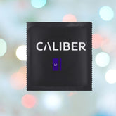 A black package labeled "Caliber" in white, featuring a purple rectangle with an "S." The backdrop displays blurred pastel lights.